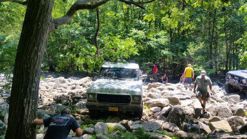 1979 Ford Bronco - A light-colored Ford Bronco navigates a rocky terrain in a wooded area, with several people observing and assisting.