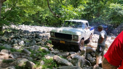 1979 Ford Bronco - A white 1970s Ford pickup truck navigates a rocky trail in a wooded area. Another dark SUV is visible further back on the trail.
