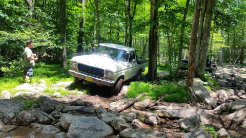 1979 Ford Bronco - A white 1970s Ford pickup truck navigates a rocky, wooded trail with a person standing nearby observing. Another off-road vehicle is visibl…