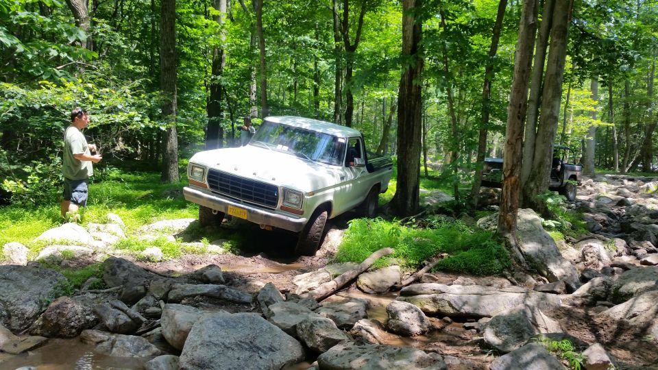 White 1970s Ford pickup truck off-roading on a rocky, wooded trail.