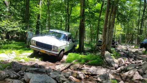 1979 Ford Bronco - A white Ford pickup truck navigates a rocky, wooded trail. Another off-road vehicle is visible further down the path.