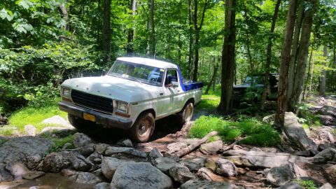1979 Ford Bronco - A white and blue 1970s Ford pickup truck navigates a rocky, wooded trail with a dark SUV visible in the background.