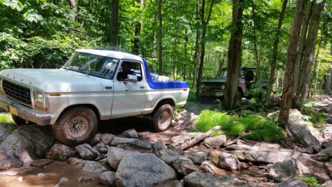 1979 Ford Bronco - A white and blue Ford Bronco is navigating a rocky, wooded trail. Another dark colored vehicle is visible behind it.