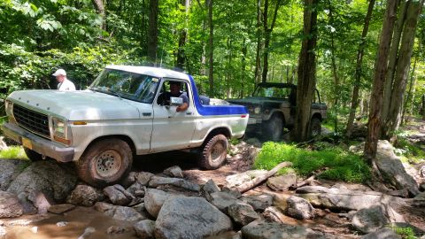 1979 Ford Bronco - A white and blue 1970s pickup truck is navigating a rocky, wooded trail, with a dark-colored SUV following behind it. The truck is angled o…