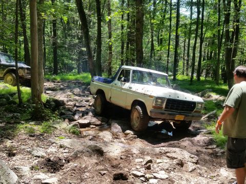 1979 Ford Bronco - A white 1970s Ford pickup truck navigates a rocky, wooded trail. Another off-road vehicle is visible in the background.