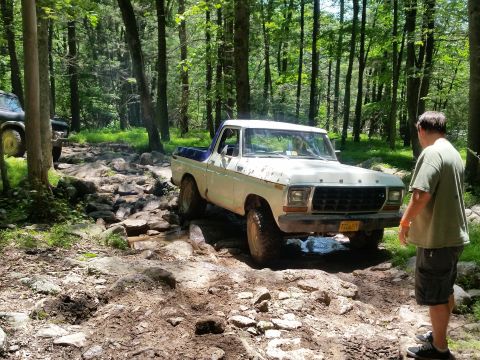 1979 Ford Bronco - A white and blue 1979 Ford Bronco is navigating a rocky, wooded trail. A man in a green shirt stands near the front of the vehicle, observi…