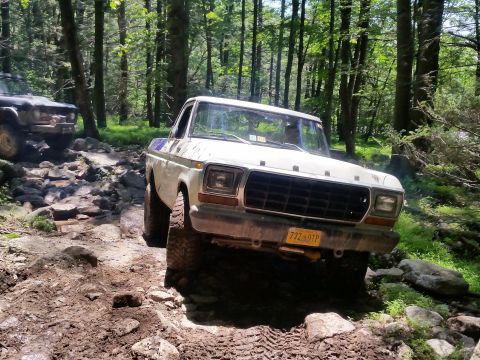 1979 Ford Bronco - A white 1970s Ford pickup truck is navigating a rocky, muddy trail in a forest. The truck is angled towards the viewer, showing its front g…