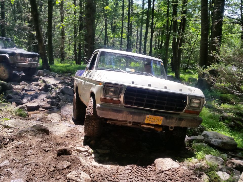 White 1970s Ford pickup truck off-roading on a muddy, rocky forest trail.