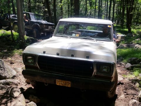 1979 Ford Bronco - A white 1979 Ford Bronco is shown off-roading on a rocky, wooded trail. Another dark-colored vehicle is visible behind it.