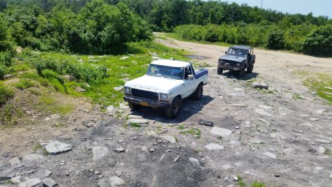 1979 Ford Bronco - Two off-road vehicles, a white Ford pickup truck and a dark-colored SUV, are parked on a rocky, dirt path surrounded by lush green trees an…