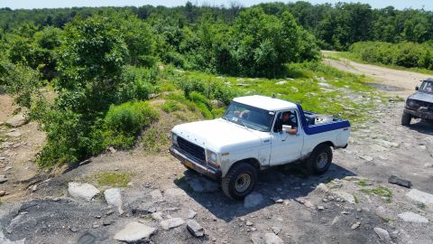 1979 Ford Bronco - A white 1979 Ford Bronco with a blue top is driving on a rocky, off-road trail with trees in the background. Another dark-colored SUV is vi…