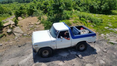 1979 Ford Bronco - A white and blue 1979 Ford Bronco is shown from a high angle, driving over a rocky, off-road terrain with trees in the background. The driv…
