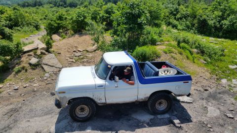 1979 Ford Bronco - A white and blue 1979 Ford Bronco is shown from a high angle, driving over a rocky, off-road trail surrounded by lush green trees.