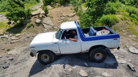 1979 Ford Bronco - A white and blue 1979 Ford Bronco is shown driving off-road on a rocky terrain with lush green trees in the background. The driver is visib…