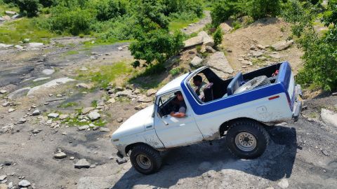 1979 Ford Bronco - A white and blue Ford Bronco is navigating a rocky, off-road trail. Two people are visible inside the open-top vehicle.