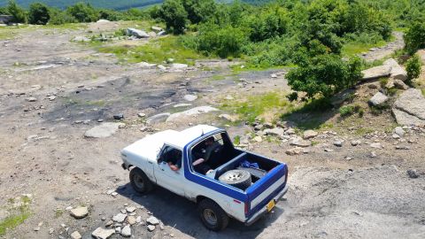 1979 Ford Bronco - A white and blue Ford Bronco is parked on a rocky, off-road trail with trees and hills in the background. The vehicle has its top removed,…