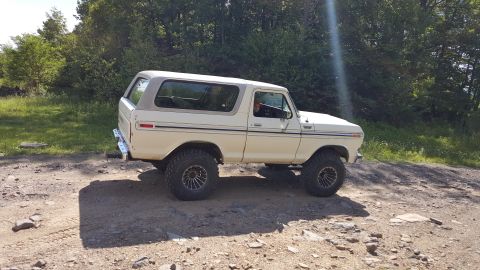 1979 Ford Bronco - A lifted, cream-colored 1979 Ford Bronco with aftermarket wheels sits on a dirt road surrounded by trees. The sun shines brightly, creating…