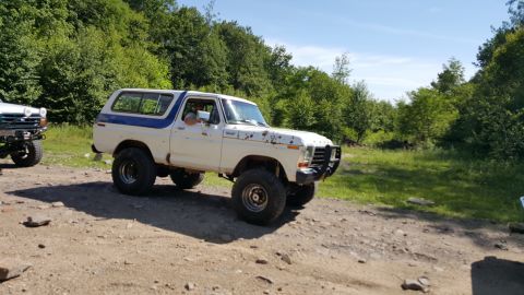 1979 Ford Bronco - A white and blue 1979 Ford Bronco with off-road tires and a brush guard is parked on a dirt road with trees in the background.