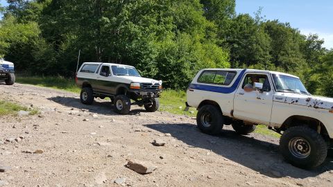 1979 Ford Bronco - Two lifted Ford Broncos are driving on a gravel road surrounded by trees. The Bronco in the foreground is white with blue accents and shows…