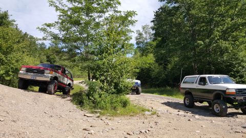 1979 Ford Bronco - Three lifted Ford Broncos are parked on a dirt road surrounded by trees. The Bronco in the foreground is white with dark green accents and…