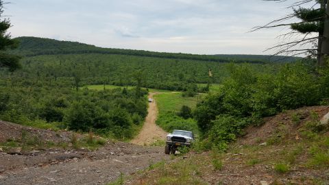 1979 Ford Bronco - A white Ford Bronco is driving down a dirt road in a wooded, hilly area. The vehicle is viewed from a high angle, looking down the slope.