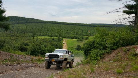 1979 Ford Bronco - A lifted, light-colored 1979 Ford Bronco drives up a dirt road with a dense forest in the background. Another white vehicle is visible furt…