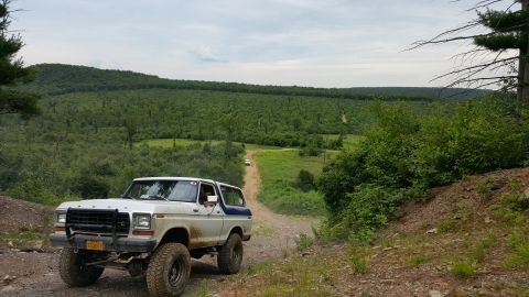 1979 Ford Bronco - A white and blue Ford Bronco is parked on a dirt road with a scenic view of a tree-covered valley and hills under a cloudy sky. Another whi…