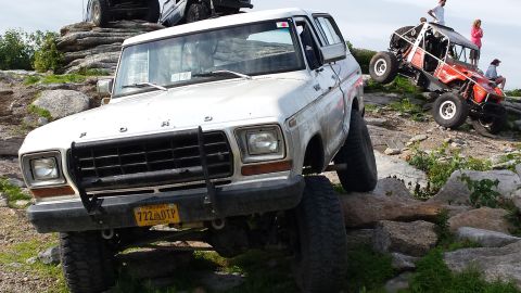 1979 Ford Bronco - A lifted white 1979 Ford Bronco is parked on a rocky incline, with a bright orange off-road buggy climbing the rocks behind it. The Bronco…