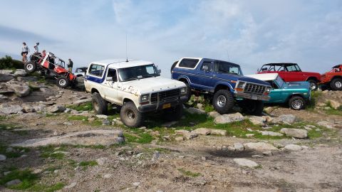 1979 Ford Bronco - Several off-road modified Ford Broncos are parked on a rocky incline under a partly cloudy sky. The vehicles are of various colors includin…