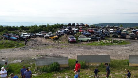1979 Ford Bronco - A large group of classic Ford Broncos and other off-road vehicles are parked on a rocky, elevated terrain with a wooded landscape in the ba…