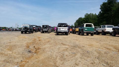 1979 Ford Bronco - A line of classic Ford Broncos and other SUVs are parked on a dirt lot under a partly cloudy sky. The vehicles are mostly facing away from…