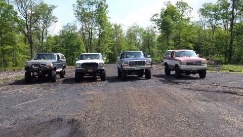 1979 Ford Bronco - Four lifted Ford Broncos are parked in a line on a dirt road with a wooded area in the background. The Broncos are various colors including…