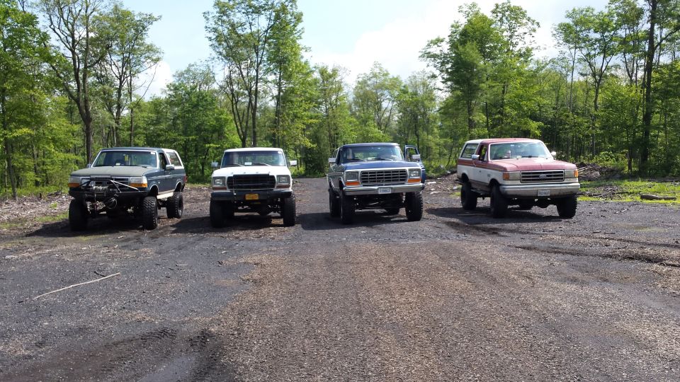 Four lifted Ford Broncos in a line on a dirt road, wooded background.