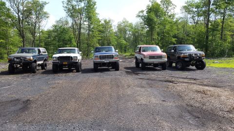 1979 Ford Bronco - Five lifted Ford Broncos and pickups are parked in a line on a gravel lot with a wooded background.