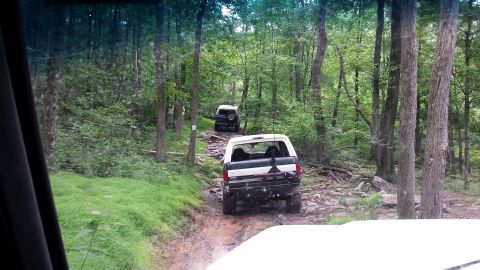 1979 Ford Bronco - Two off-road vehicles are navigating a rocky, muddy trail through a dense forest. The lead vehicle, a white and green SUV, is seen from the…