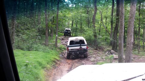 1979 Ford Bronco - Two off-road vehicles navigate a rocky, muddy trail through a dense forest. The lead vehicle, a white and green SUV, is seen from the rear,…