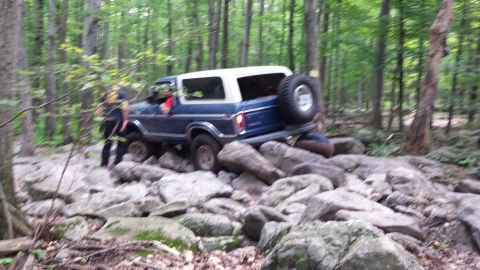 1979 Ford Bronco - A blue and white Ford Bronco is navigating a rocky trail in a forest. The vehicle is angled to the right, showing its rear and side profile…
