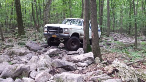 1979 Ford Bronco - A white 1970s Ford pickup truck with a black brush guard is navigating a rocky, wooded trail. The truck is positioned at an angle, with its…