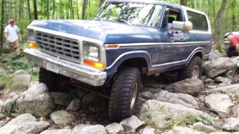 1979 Ford Bronco - A blue 1979 Ford Bronco is navigating a rocky, wooded trail. The vehicle is angled to the right, with its front tire clearly visible on a l…