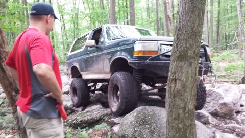1979 Ford Bronco - A dark green Ford Bronco is navigating a rocky, wooded trail. A person wearing a red shirt and a baseball cap stands to the left of the veh…