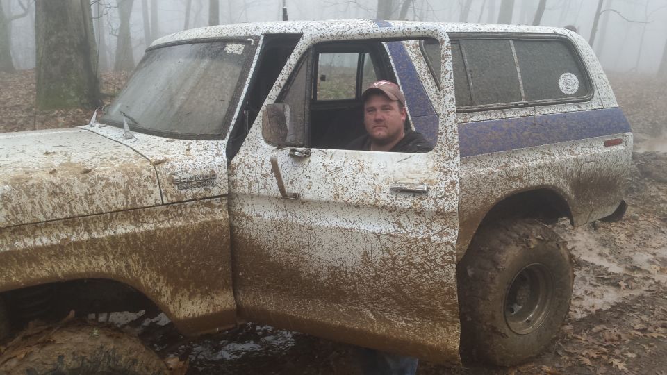 Mud-covered white 1979 Ford Bronco with a man in the driver's seat in a foggy forest.