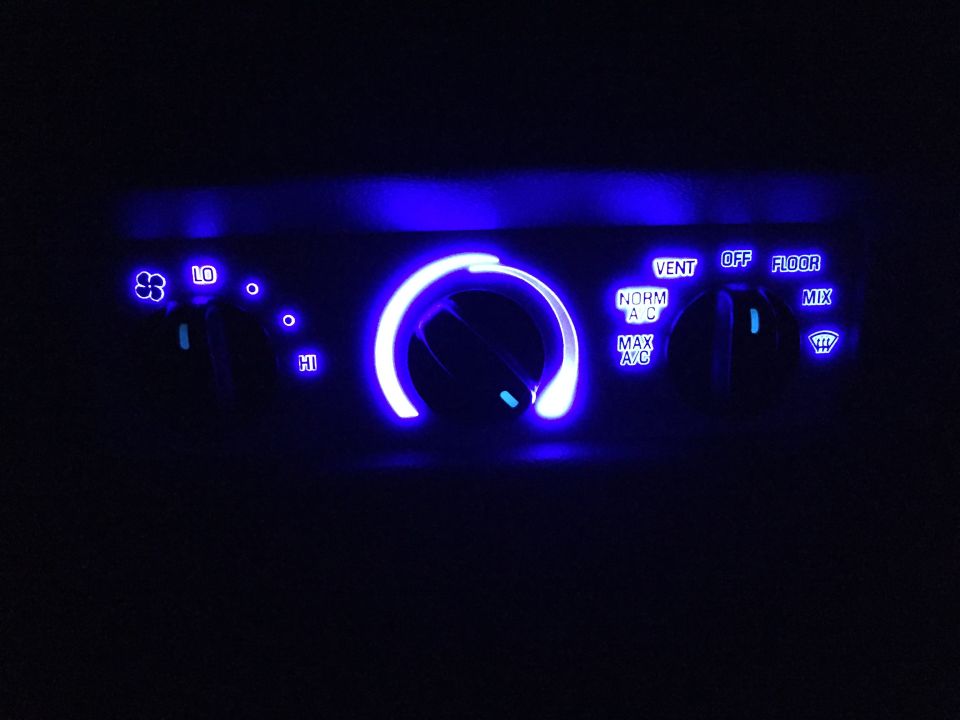 Car climate control panel with blue illuminated knobs and labels for fan speed and air direction.