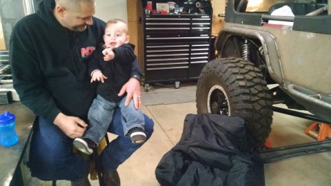 1985 Jeep CJ7 - A man holds a baby in a garage in front of a modified Jeep CJ7. The Jeep has large off-road tires and is supported by jack stands.