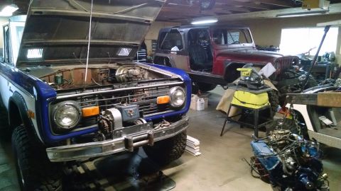 1985 Jeep CJ7 - Two off-road vehicles are in a garage with their hoods open. A blue and white Ford Bronco is in the foreground, and a red Jeep CJ is in the…