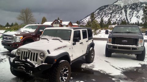 1985 Jeep CJ7 - A white Jeep Wrangler Rubicon is parked in the snow next to two Ford pickup trucks. A snow-covered mountain and houses are visible in the b…