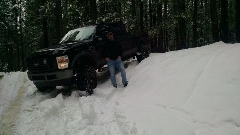 1985 Jeep CJ7 - A man poses next to a black Ford F-Series pickup truck stuck in deep snow on a wooded trail.