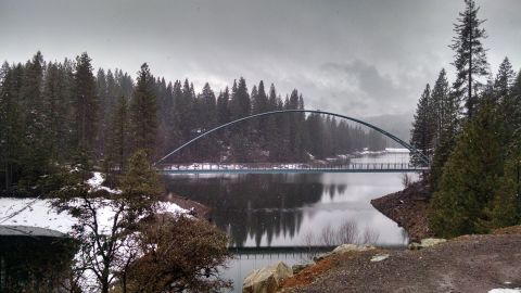 1985 Jeep CJ7 - A scenic view of a bridge arching over a calm body of water, surrounded by snow-dusted pine trees under a cloudy sky.