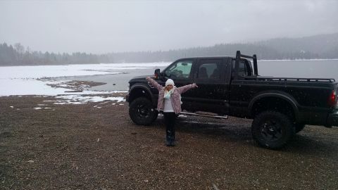 1985 Jeep CJ7 - A woman stands with her arms outstretched in front of a black pickup truck parked on a gravel shore next to a body of water. Snow is fallin…