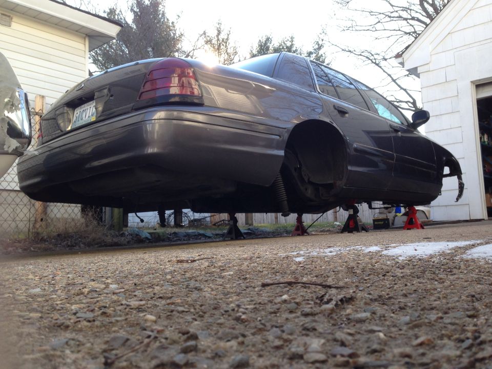 Dark gray sedan on jack stands, rear view, front fender and bumper missing.