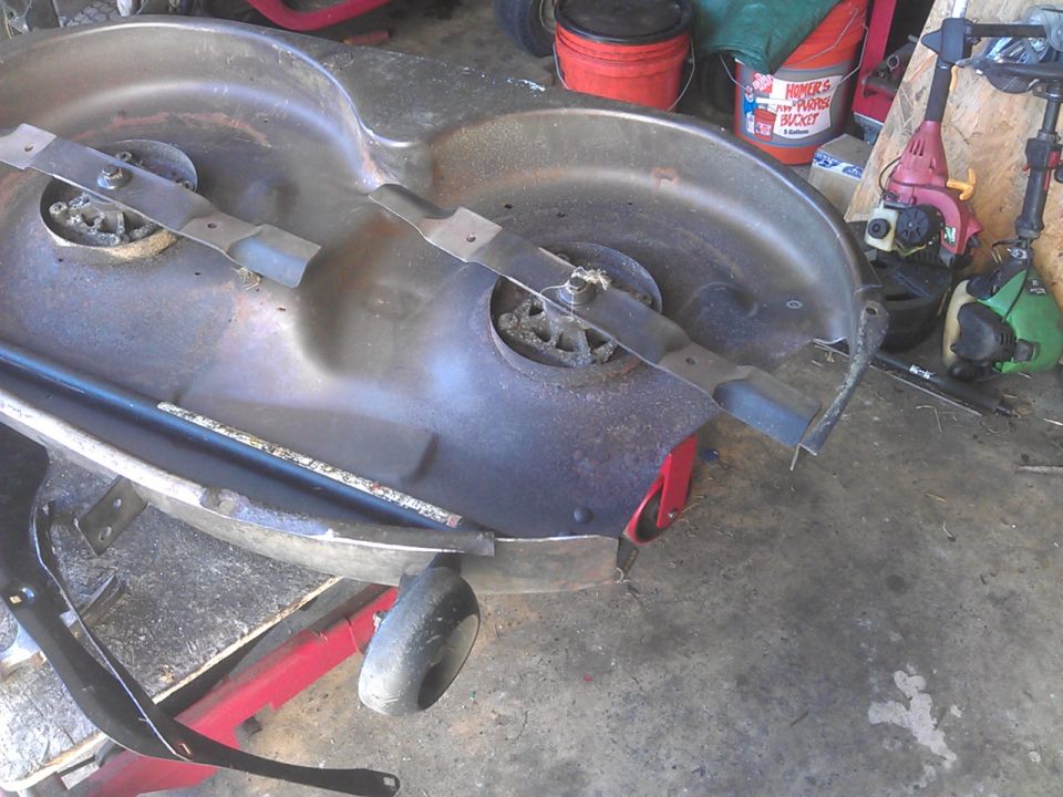 Underside of a rusty lawnmower deck with blades, tools in background.